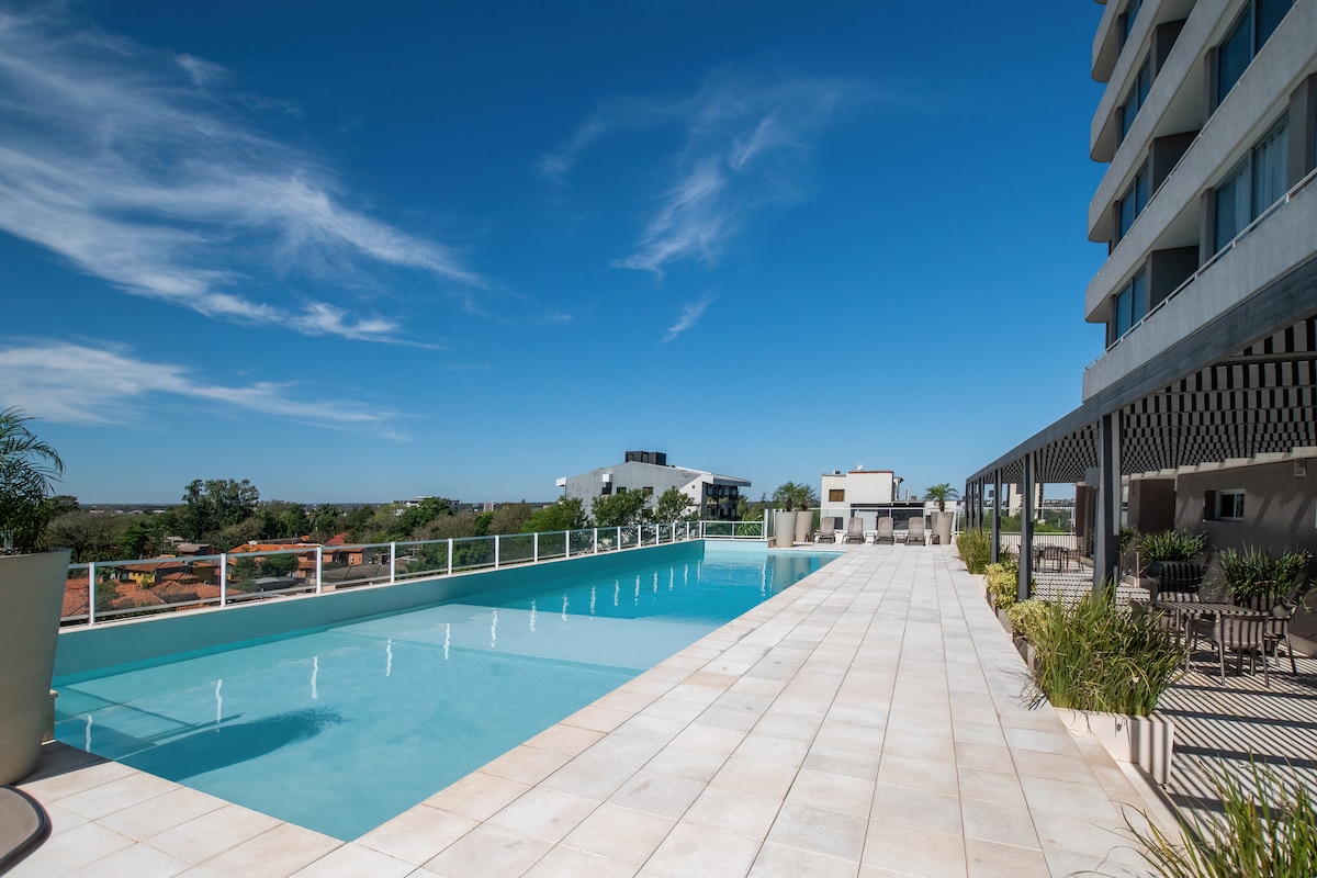 An elongated swimming pool reflects the clear blue sky, bordered by a well-maintained deck featuring potted plants. Comfortable lounge chairs are arranged along the poolside, providing a relaxing space for guests. The modern building is visible in the background, enhancing the tranquil ambiance.