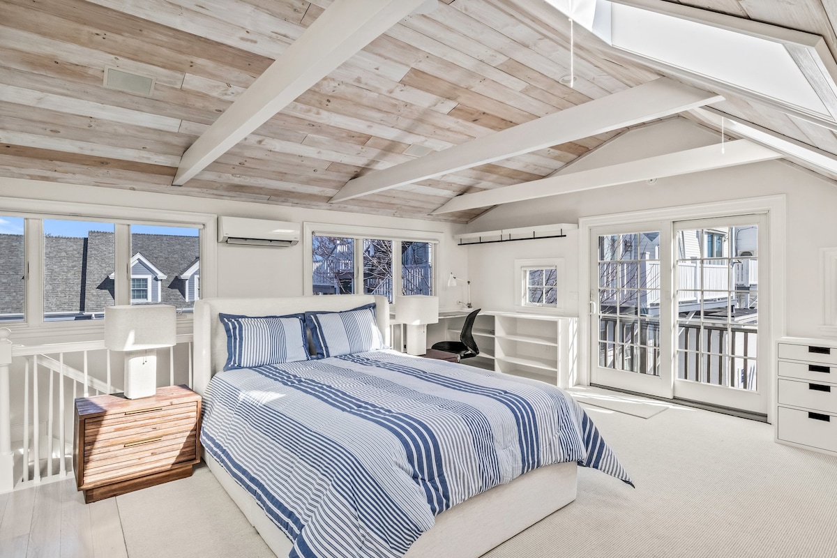 An open-concept bedroom suite is highlighted by a spacious bed adorned with blue and white striped bedding. Natural light filters through three skylights and multiple windows, illuminating the crisp white walls and wooden ceiling beams. Access to a private balcony is available through sliding glass doors.