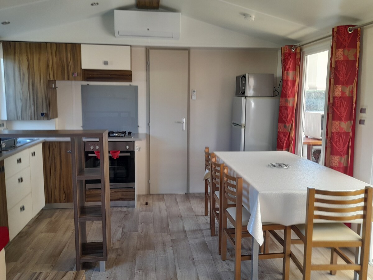 An open kitchen area features modern cabinetry and appliances, including a refrigerator and gas stove. A dining table with six wooden chairs sits under a bright window draped with red curtains, allowing natural light to fill the space.
