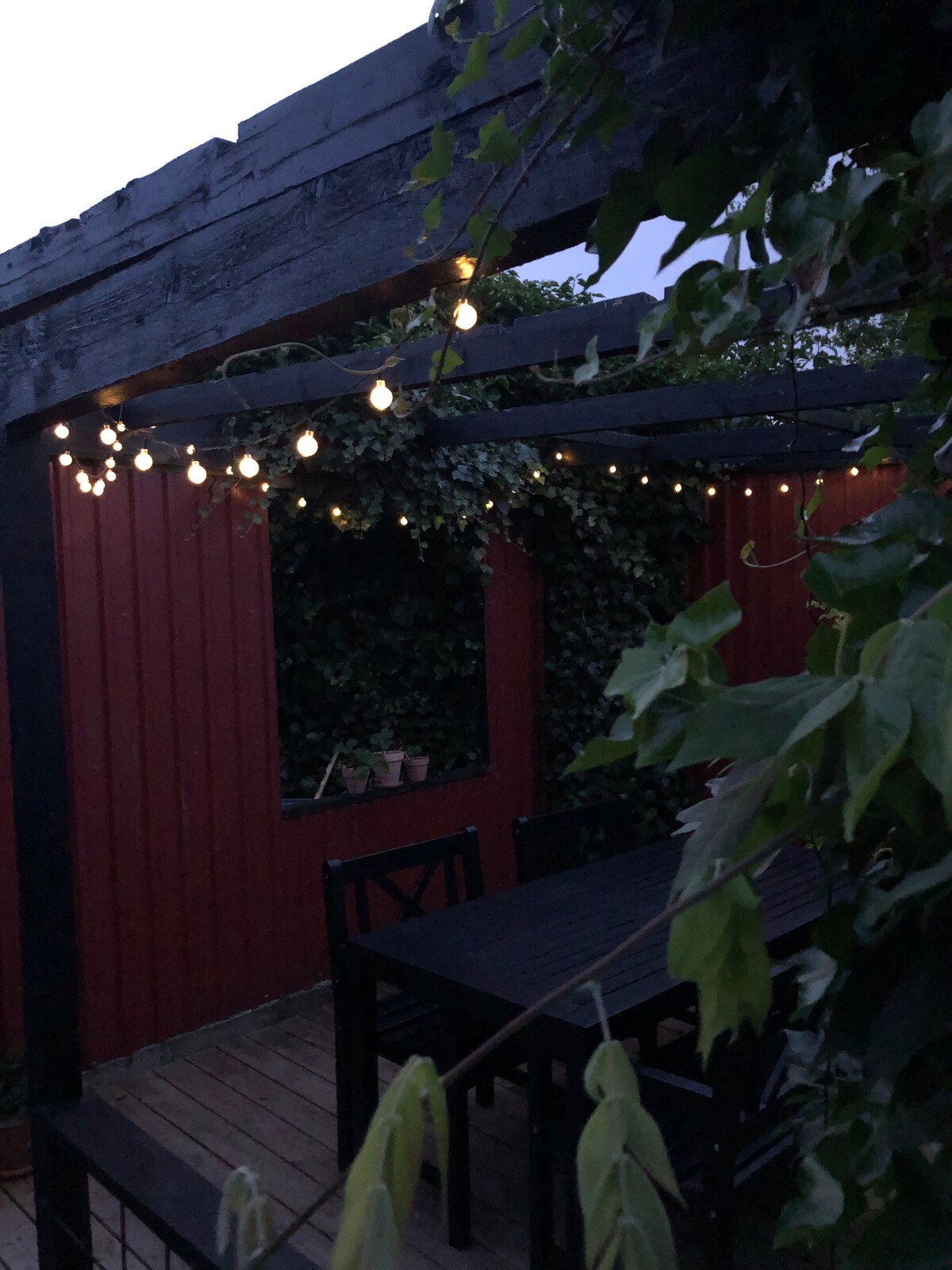 A cozy outdoor terrace features a dark wooden table and chairs, surrounded by lush greenery. Soft string lights hang overhead, creating a subtle ambiance. The red wooden walls provide a contrasting backdrop, enhancing the inviting outdoor space.