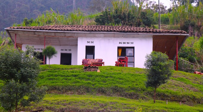 A cottage is set on a gently sloping green hillside, featuring a traditional tiled roof and white walls. Large windows offer scenic views of the surrounding landscape, while a small terrace is furnished with seating, providing a welcoming outdoor space for relaxation.