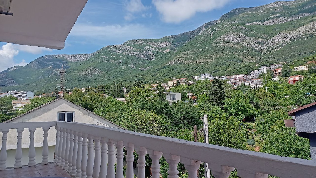 A spacious terrace features white balustrades overlooking lush greenery and distant mountains. The landscape includes houses nestled among trees, with a blue sky and soft clouds above, creating a serene outdoor environment.