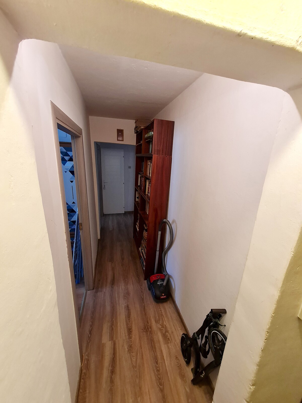 A narrow hallway is shown, featuring light-colored walls and a wooden floor. A bookshelf filled with books stands against one side, while a vacuum cleaner and a folding stroller are located near the entrance. A doorway at the end leads into another room.