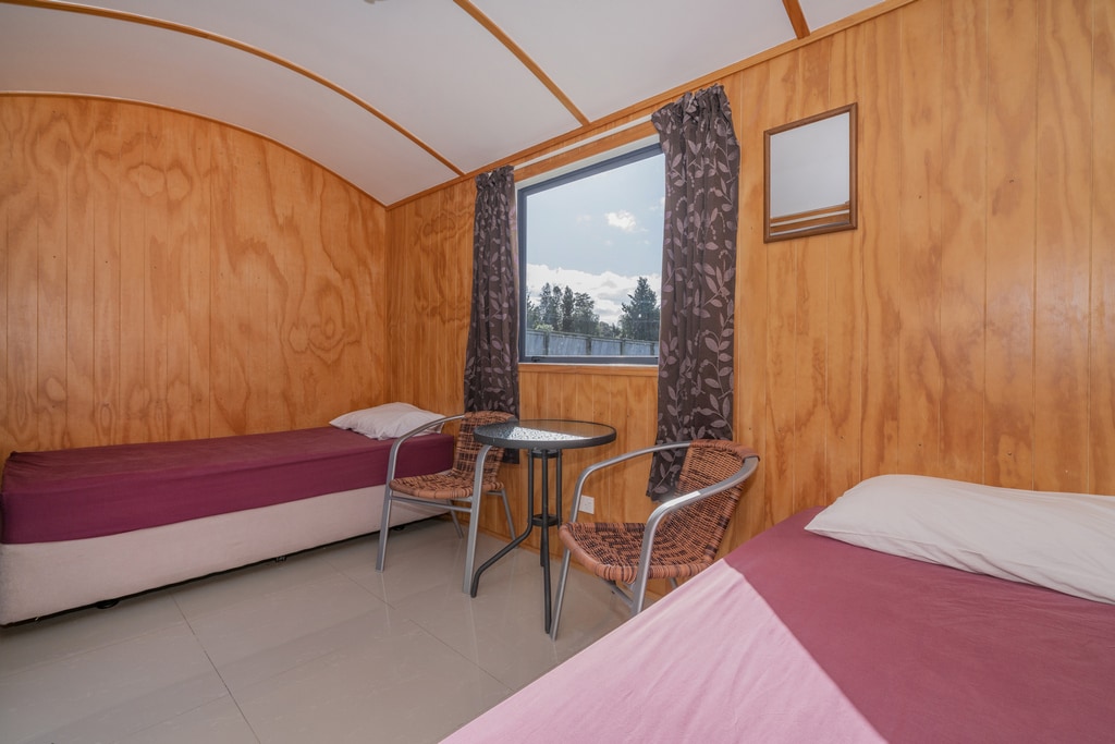 An interior space features two single beds with fitted sheets, positioned near a window with floral drapes. A small round table and two chairs sit between the beds, offering a cozy spot for conversation. Light streams through the window, illuminating the wooden walls.