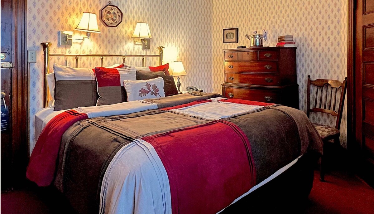 A cozy bedroom features a large bed adorned with decorative pillows and a blanket in rich tones of red and brown. Beside the bed, wall sconces provide soft lighting. A wooden dresser and a chair are positioned against the patterned wallpaper.