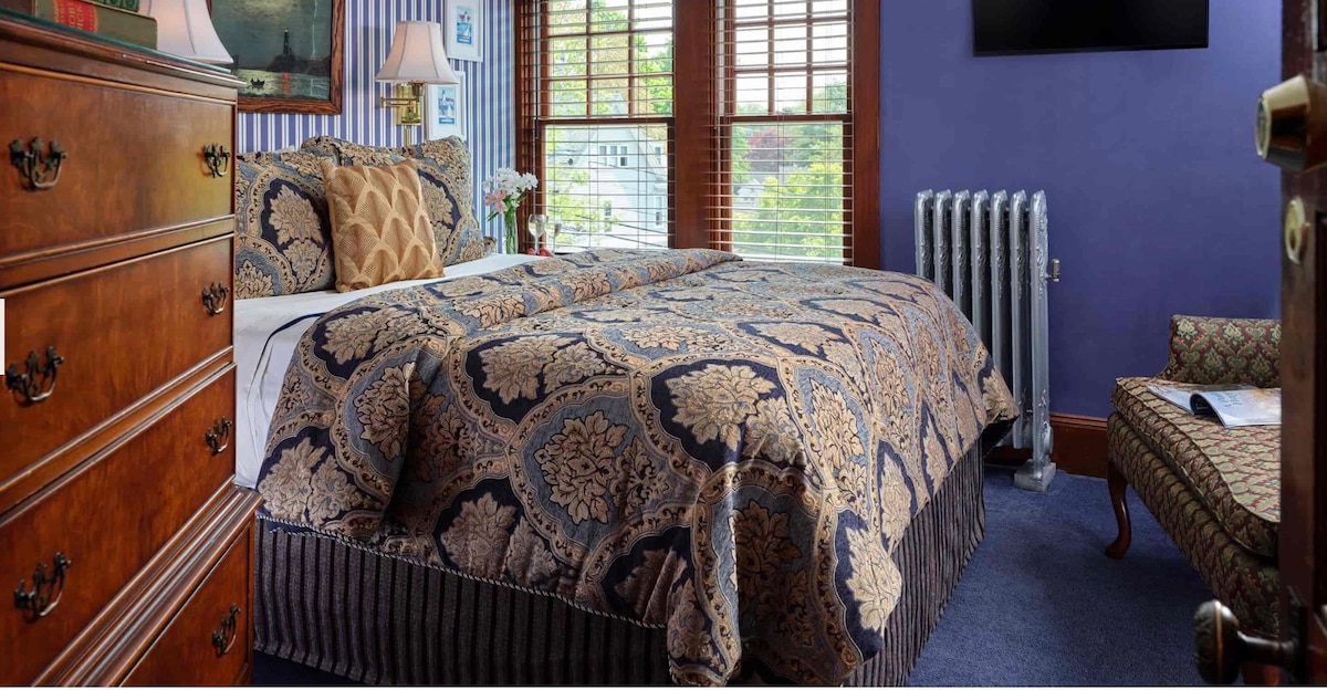 A comfortable bedroom features a patterned duvet covering a spacious bed with accent pillows. Natural light filters through wooden blinds, illuminating the room. A vintage dresser and a cozy seating area with a patterned chair add charm, while a radiator provides warmth.