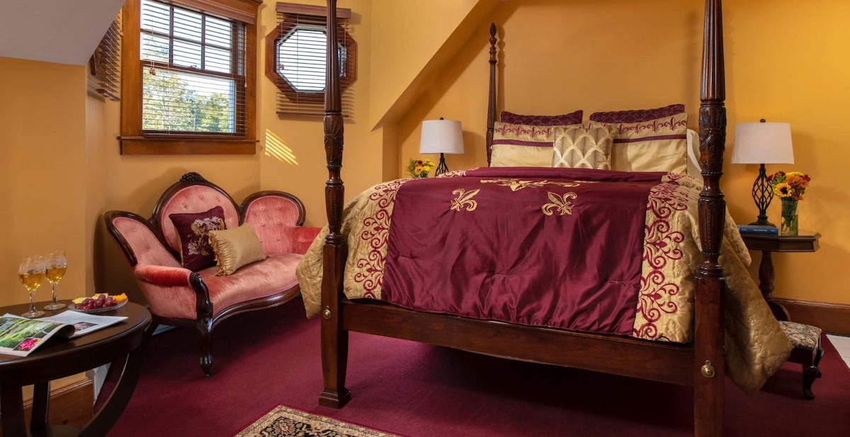 A traditional high bed with a carved mahogany frame is adorned with a rich burgundy coverlet and decorative pillows. A cozy sitting area features a pink loveseat next to a small table with refreshments. Natural light streams through a bay window, illuminating the warm yellow walls.