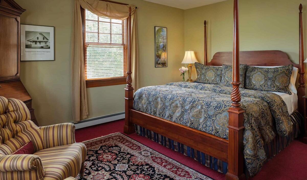 A spacious room features a carved cherry four-poster bed adorned with intricate bedding. Natural light filters through wooden blinds, illuminating the surroundings. A comfortable armchair sits beside a decorative rug, and artwork complements the neutral wall color.