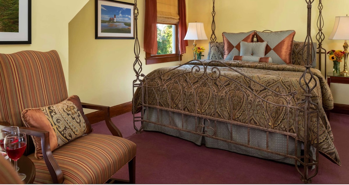 A softly-lit room features a spacious bed framed by ornate ironwork, complemented by decorative pillows. A seating area presents a striped armchair next to a small table with two glasses of red beverage. Windows allow natural light to illuminate warm, yellow walls.