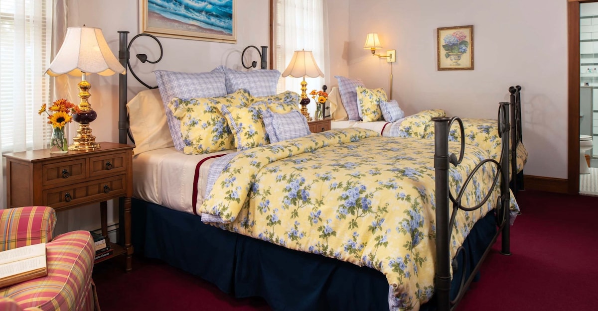 A spacious room features a queen bed and a twin bed adorned with floral-patterned bedding. Two bedside lamps provide warm lighting alongside wooden nightstands, while a cozy chair and a window with sheer curtains allow natural light to brighten the space.