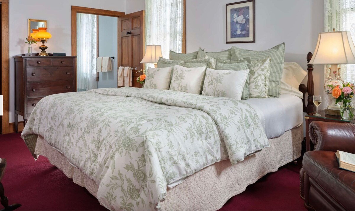 The room features a king-size bed dressed in a floral green and white comforter, surrounded by multiple pillows. A wooden dresser and two bedside lamps provide soft lighting. Natural light filters through sheer curtains, enhancing the inviting atmosphere of the space.