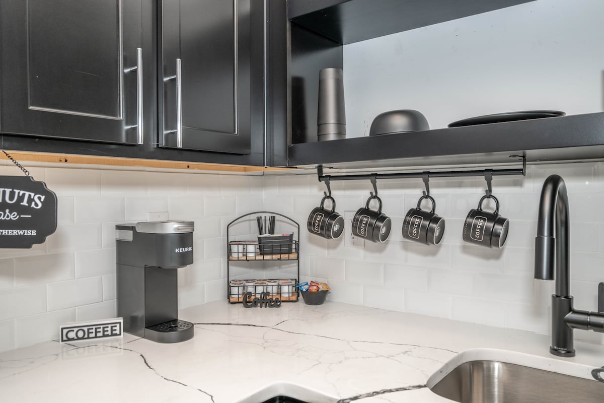 A modern kitchen area features dark cabinetry and a light countertop. A coffee maker sits beside a decorative sign, while a rack displays five coffee mugs. A small organizer holds utensils and pencils, contributing to the functional layout.