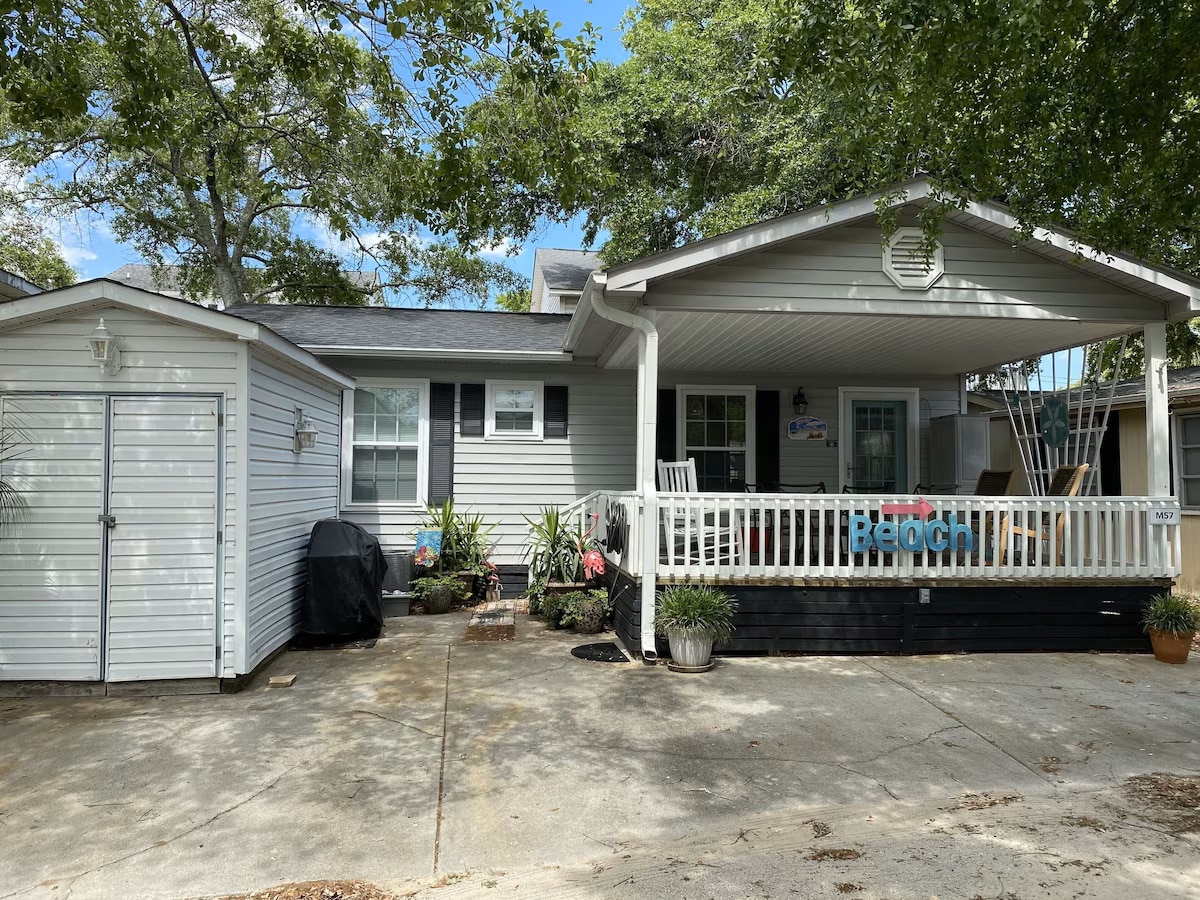 The exterior of a beach house is shown, featuring a covered porch with seating and a colorful beach sign. A nearby grill and various potted plants are visible, along with a storage shed on the left. Shade is provided by mature trees in the vicinity.