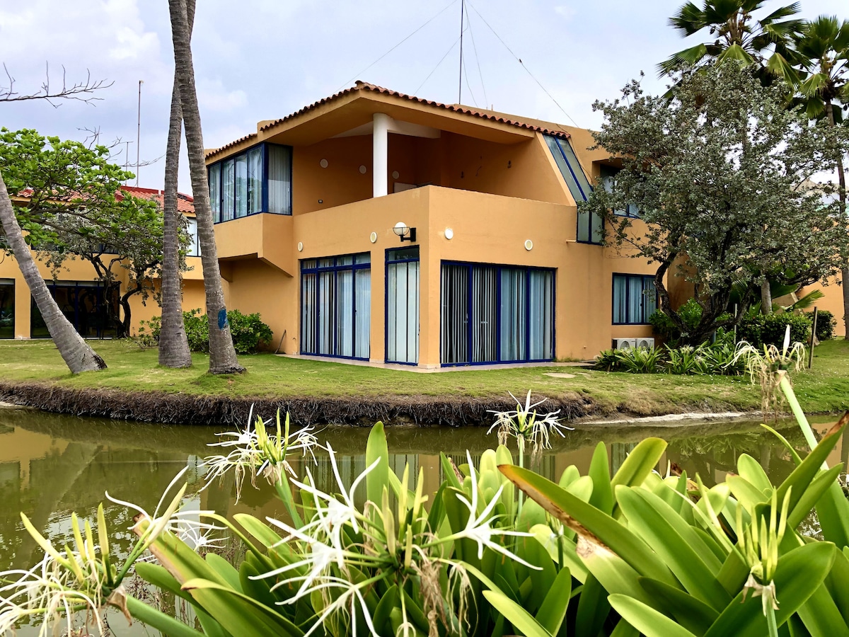 The two-story townhouse features large glass windows and doors, surrounded by lush greenery and a serene canal. A well-maintained exterior showcases a blend of vibrant colors, with palm trees and tropical plants enhancing the tranquil setting.