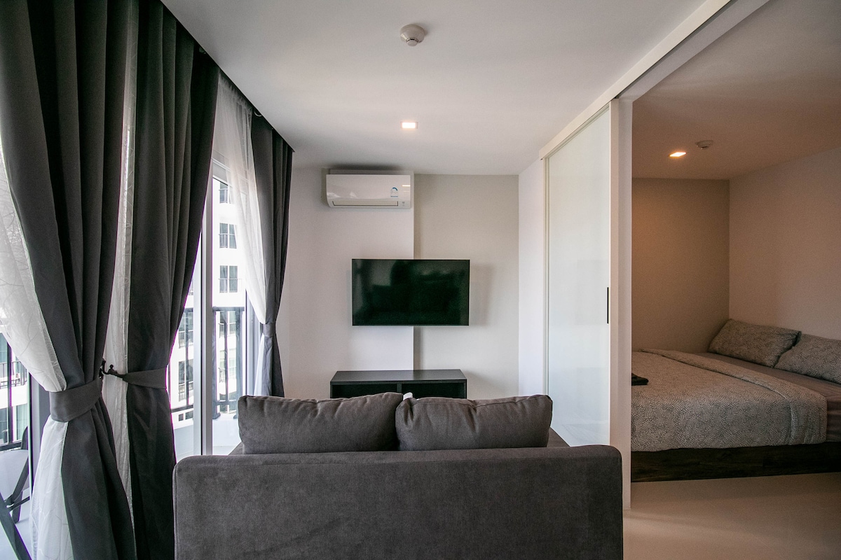 A living space is featured with a gray sofa facing a flat-screen TV mounted on the wall. Light streams in through large windows with sheer curtains, revealing a separate bedroom area with a neatly made bed in the background.