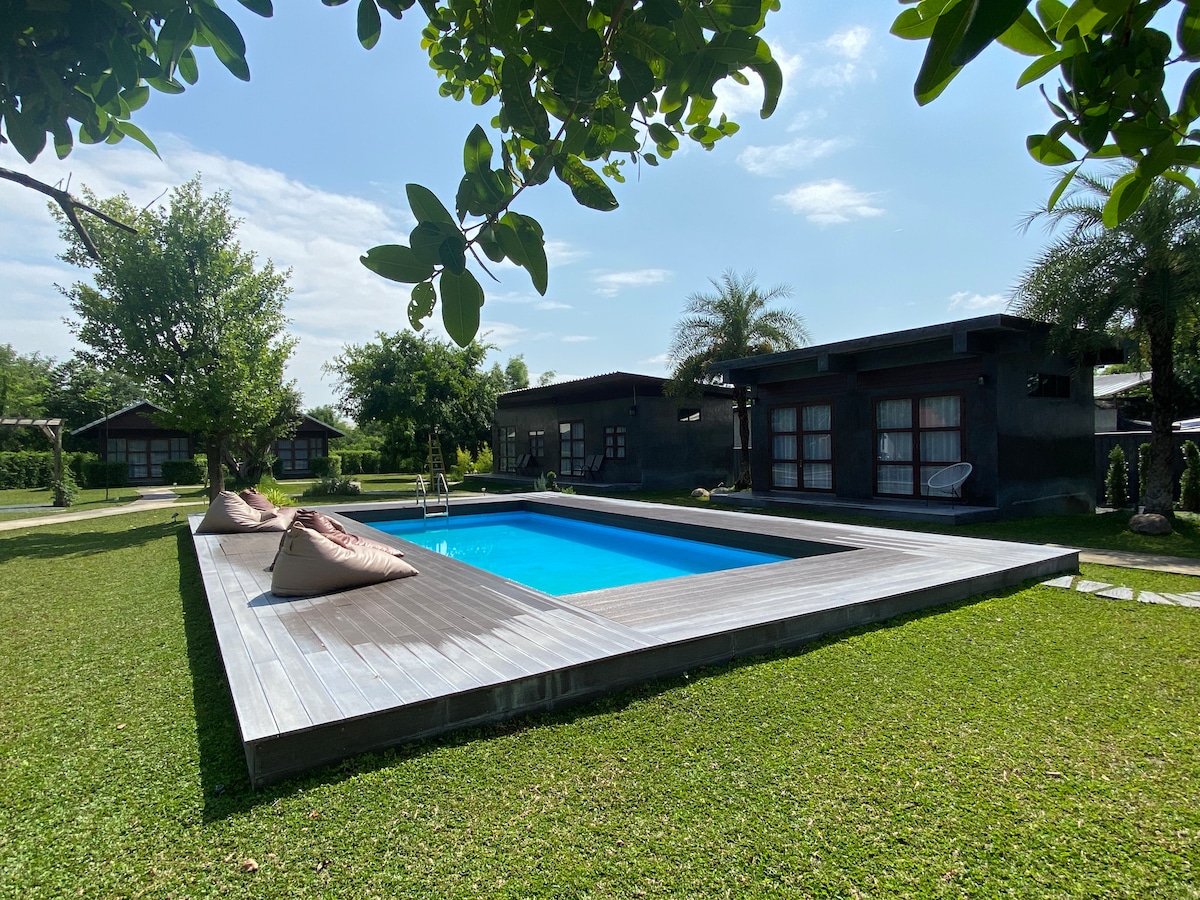 A serene swimming pool is set within a wooden deck, surrounded by lush green grass and trees. Sun loungers are arranged near the poolside, while charming cabins are visible in the background under a clear blue sky.