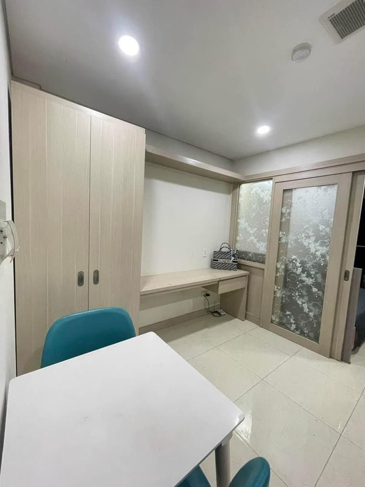 A neutral-toned room is equipped with a small white table and two turquoise chairs. Built-in cabinets and a partial glass door add to the functionality of the space, while soft overhead lighting illuminates the polished floor.