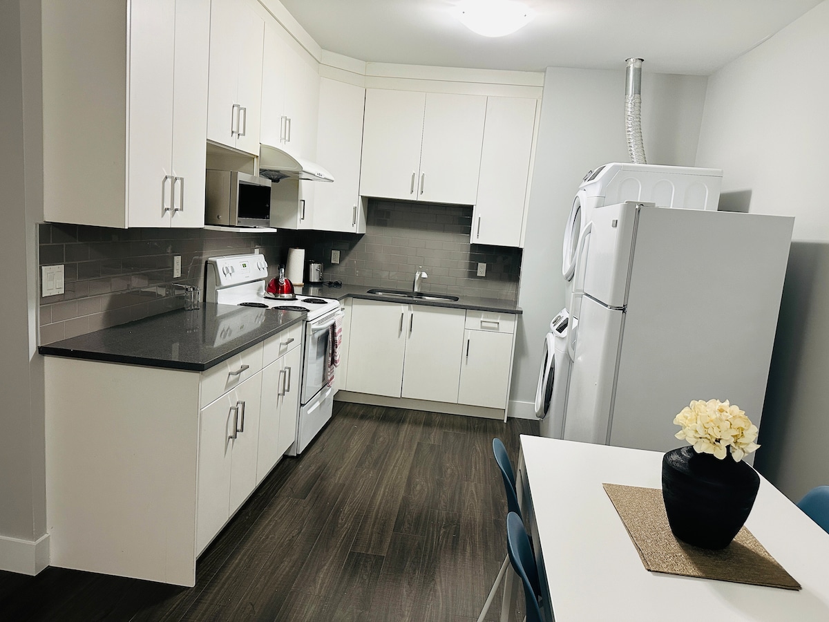 A modern kitchen is equipped with sleek white cabinetry and stainless steel appliances. A light gray backsplash complements the granite countertops. A dining table for four is present, accented by a simple vase holding white flowers, while a laundry area is visible behind the main appliances.