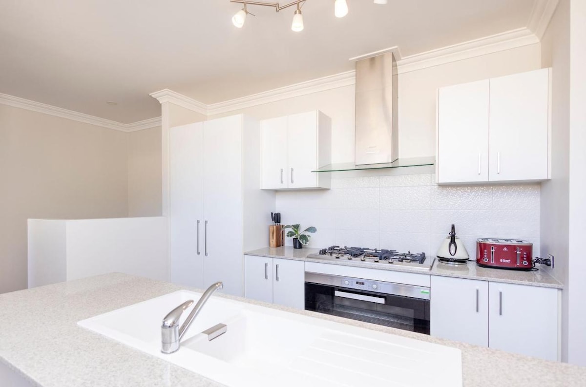 A modern kitchen features sleek white cabinetry with minimalistic handles. Stainless steel appliances, including a gas stove and a kettle, are neatly arranged. A large countertop provides ample space for preparation, complemented by an undermount sink and simple light fixtures overhead.