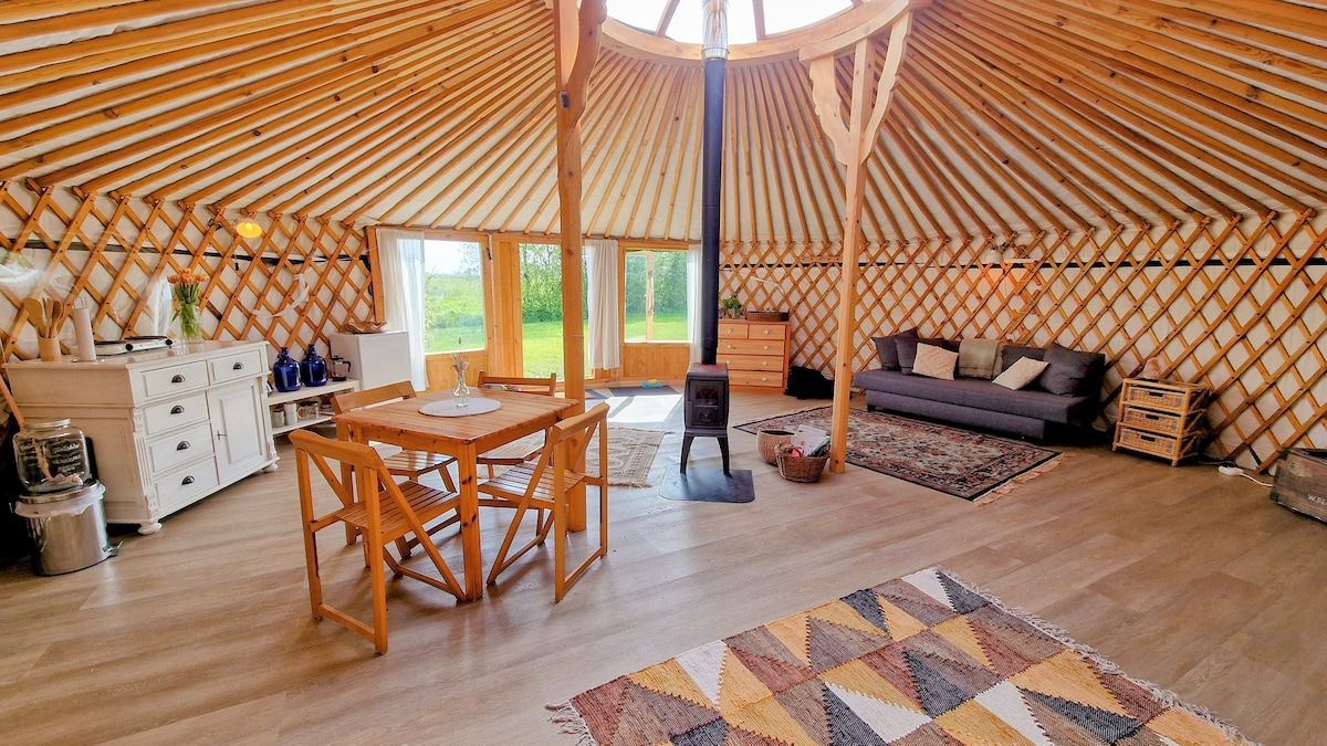 The interior of the yurt reveals a spacious layout with circular wooden beams and a central chimney. A cozy seating area features a dark sofa and a small table surrounded by wooden chairs. Natural light floods the space through a large skylight, enhancing the warmth of the wooden elements.