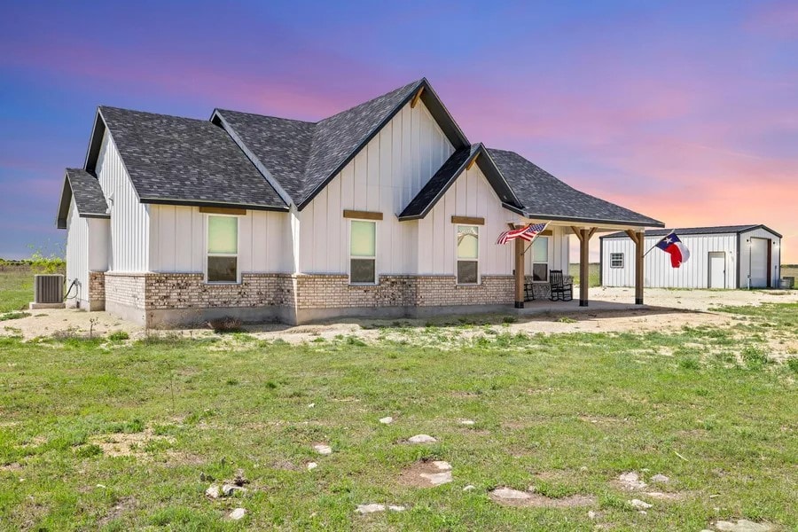 A modern ranch-style house is set against a colorful sunset sky, featuring a combination of white siding and brick accents. Two flags are displayed near the entryway. A spacious lawn surrounds the home, with a large outbuilding visible in the background.