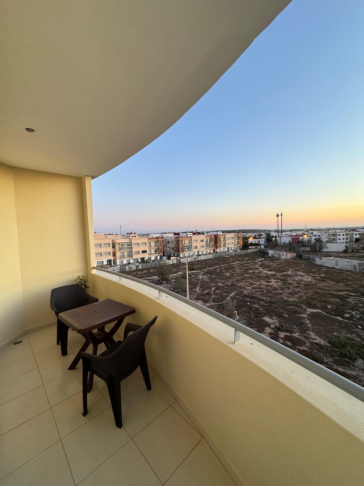 A private balcony space features a small table and two chairs, with a clear view of the surrounding neighborhood. The scene is illuminated by the soft glow of sunset, highlighting nearby buildings and open land.