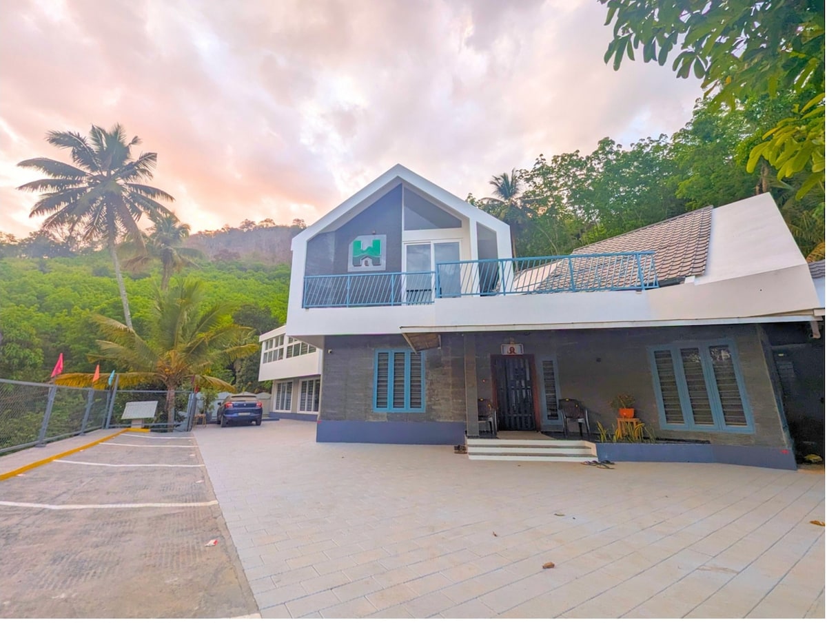 The exterior of a modern homestay is shown, surrounded by lush greenery. A two-story building features a sloped roof and a terrace with a blue railing. Parking is available in front, with palm trees framing the scene and a warm sky in the background.