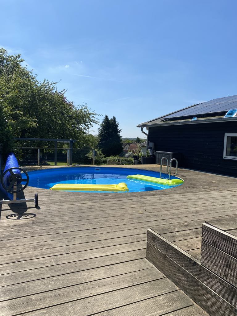 An outdoor area features a heated pool surrounded by a wooden deck. A smooth surface is seen for sunbathing and relaxation, with green trees providing shade in the background. Steps lead out of the pool, enhancing accessibility.