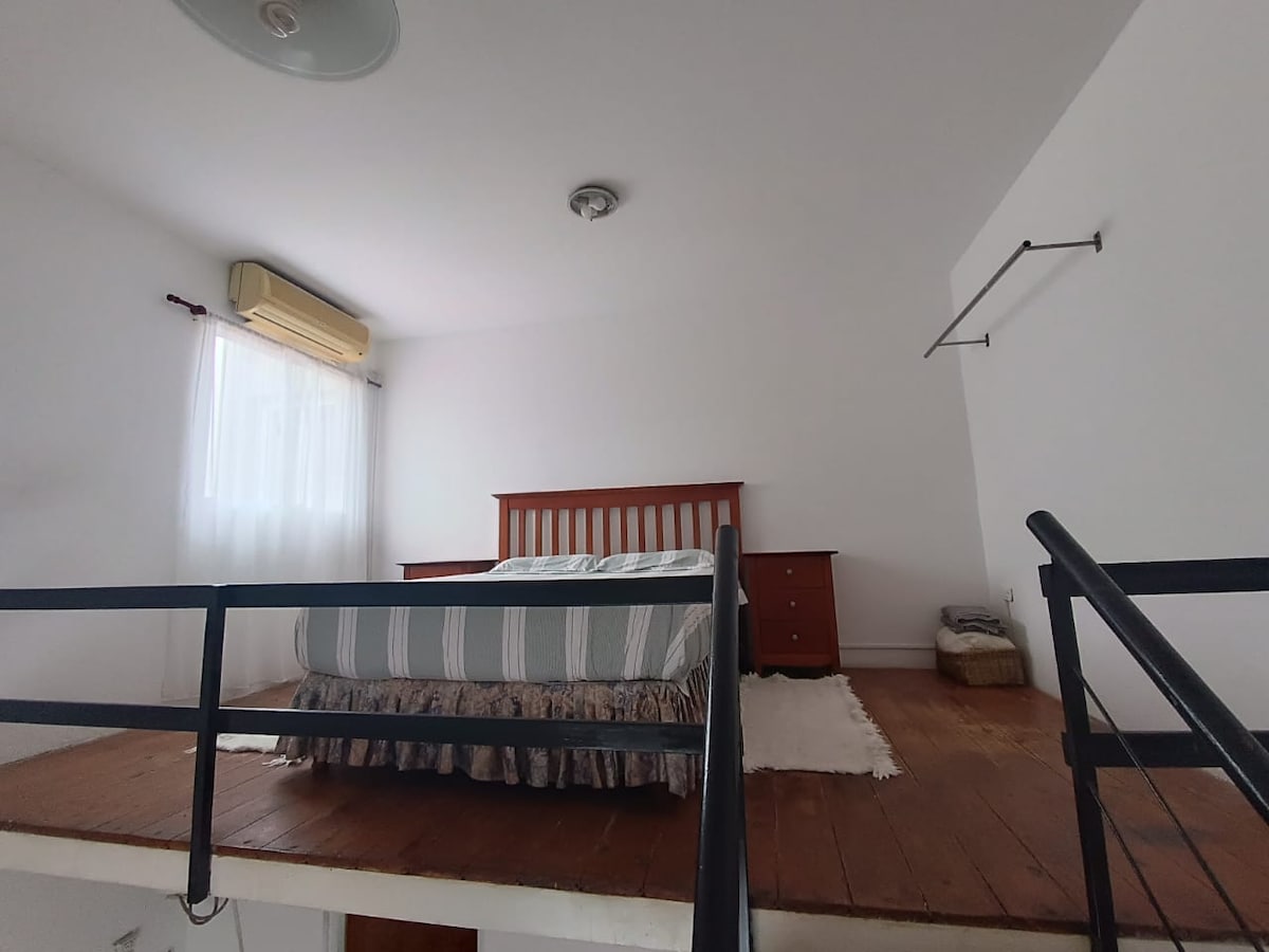 A lofted sleeping area is presented with a spacious bed, complemented by a wooden headboard and matching nightstands. Soft natural light filters through sheer curtains, illuminating the warm wood flooring. A minimalist design enhances the serene environment, creating a calm retreat feel.