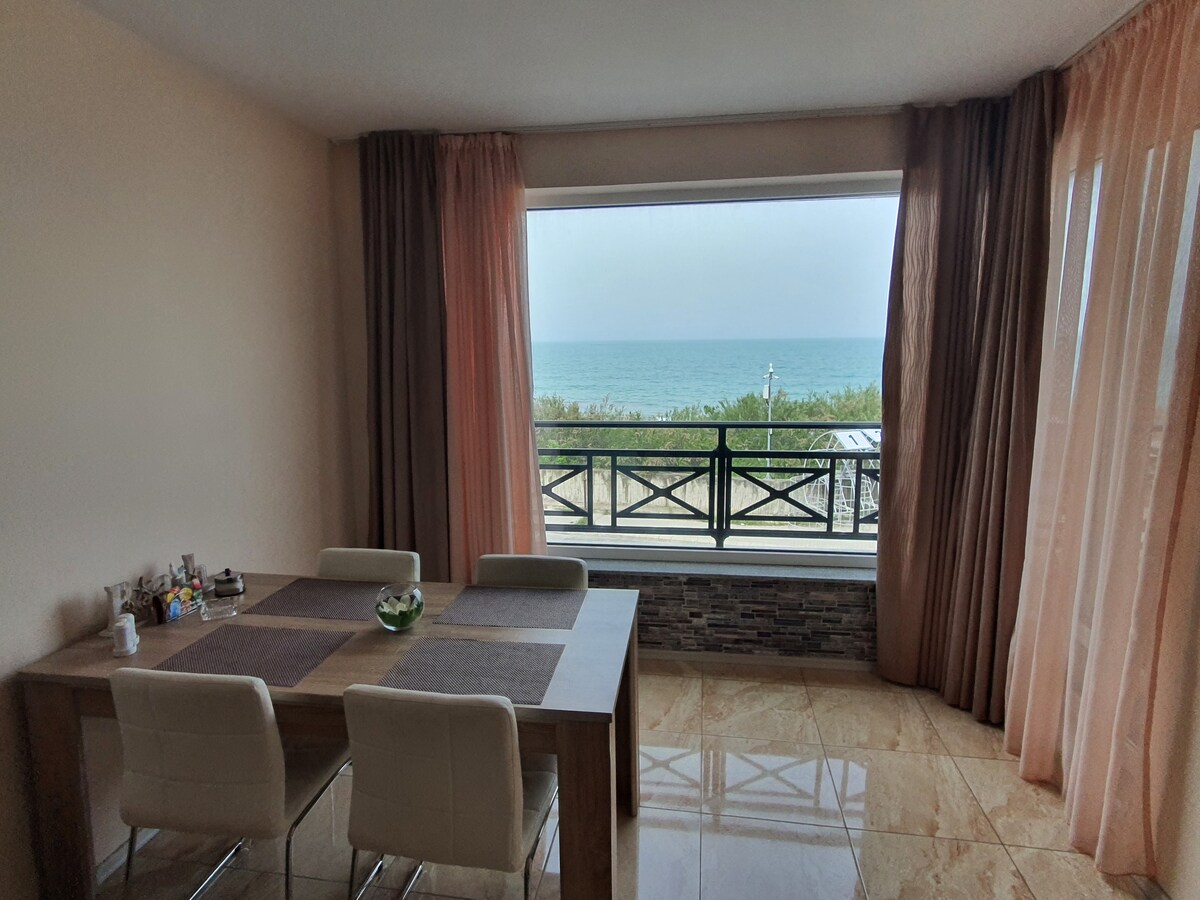 A dining area features a wooden table surrounded by four white chairs, positioned near a large window. The window offers a view of the sea, with soft drapes framing the scene. Light-colored walls complement the polished stone floor, adding to the space's brightness.