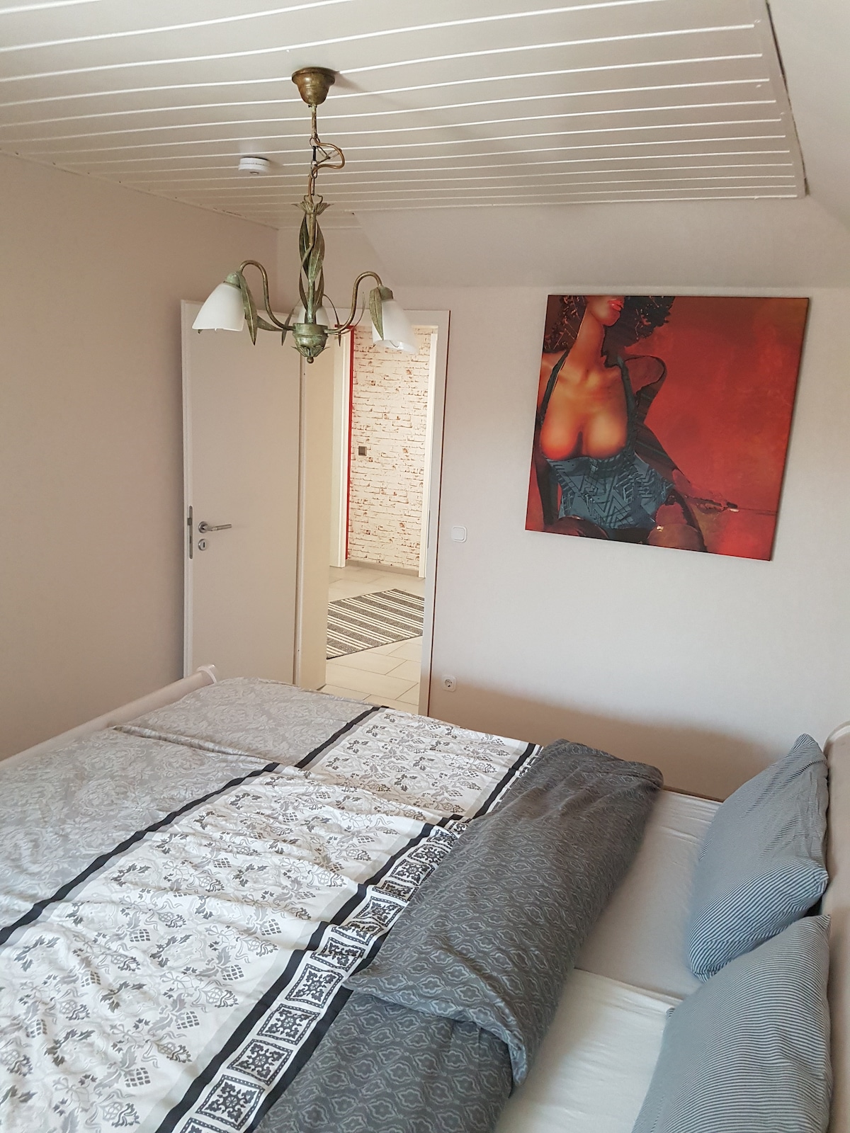 A bedroom is furnished with a spacious bed dressed in neutral tones and patterned bedding. A piece of art featuring a figure hangs on the wall above the bed, and a doorway leads to another room with a bright tiled floor.