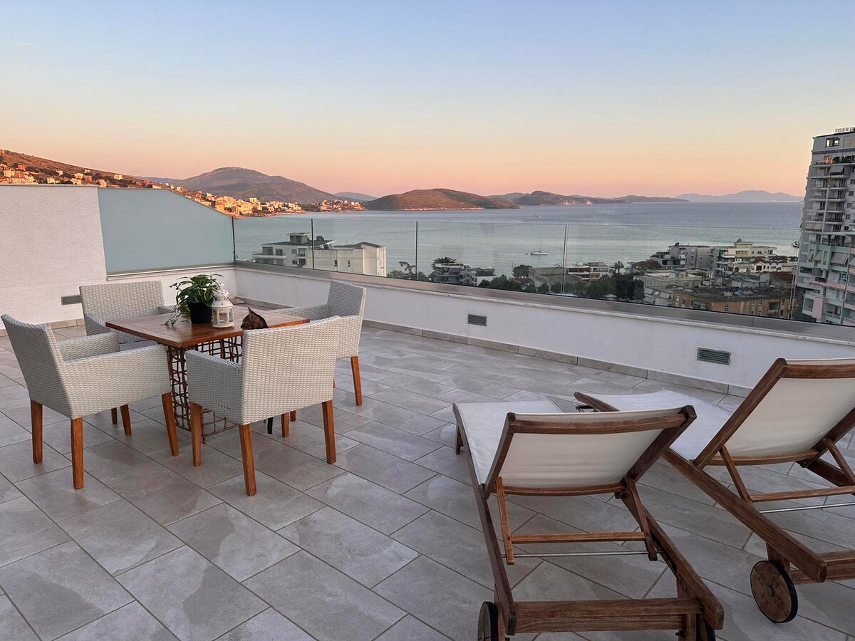 A spacious terrace is presented, featuring a wooden dining table surrounded by four wicker chairs. Two loungers are positioned nearby, offering a relaxing space. The view includes distant hills and water, with soft colors in the sky reflecting the time of day.