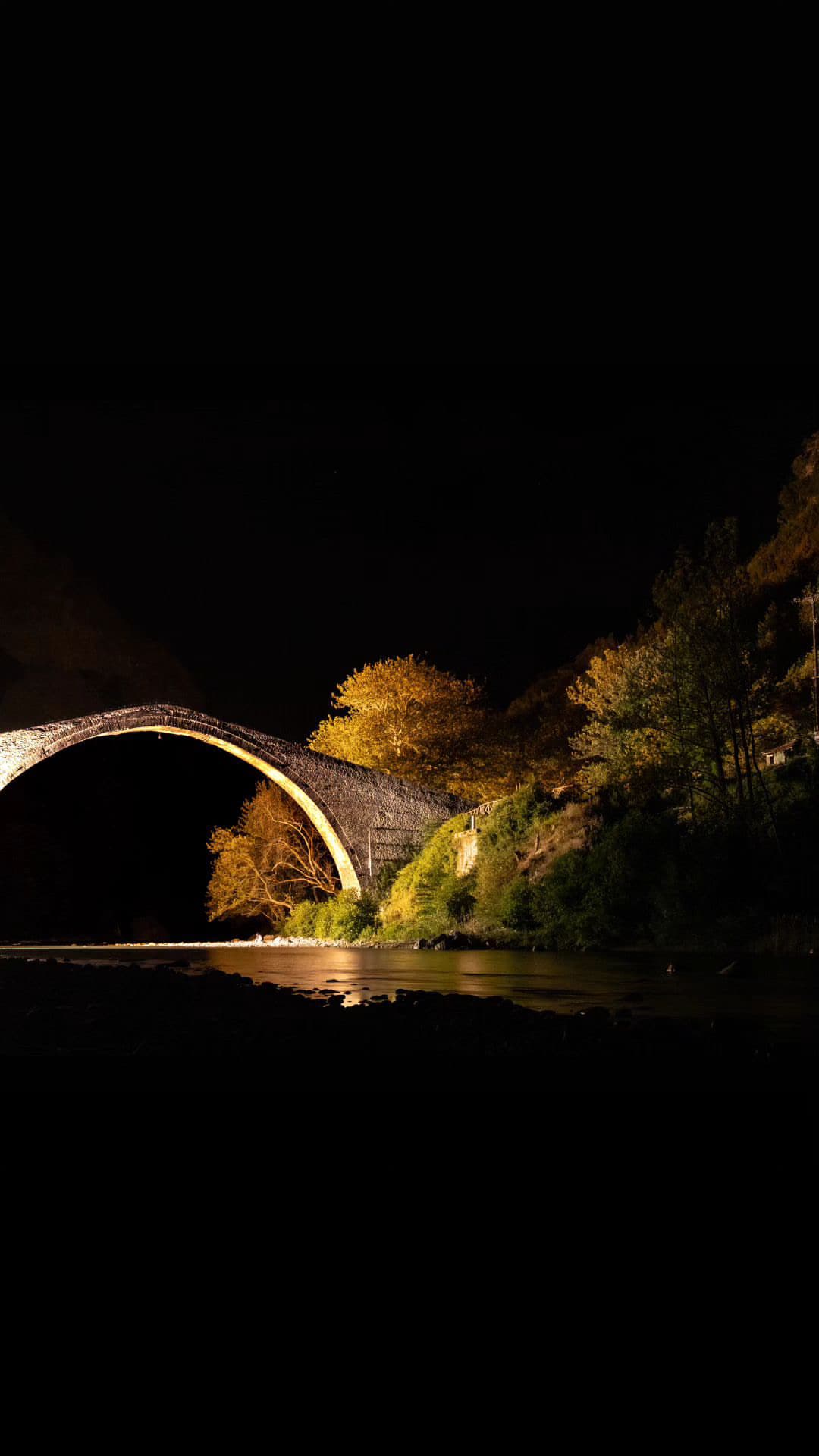 A stone bridge is illuminated at night, showcasing its arch against a dark sky. Trees with golden foliage are visible beside the water, which reflects the soft light, creating a serene and peaceful atmosphere.