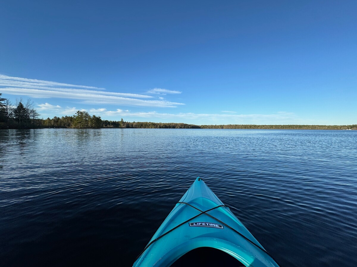 A clear blue sky is reflected on the calm waters of Baxter Lake, viewed from the front of a kayak. The shoreline features lush greenery with a mix of trees, creating a serene scene for water activities.