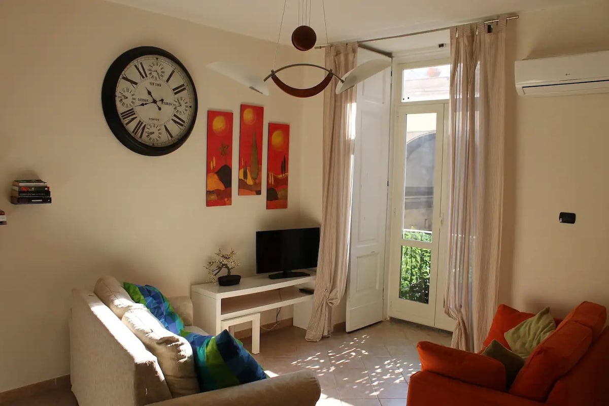 A living area features a light-colored sofa alongside an orange armchair. A television is placed on a low table, while two decorative paintings in warm tones adorn the wall. Natural light enters through a door leading to a small outdoor space, complemented by sheer curtains.