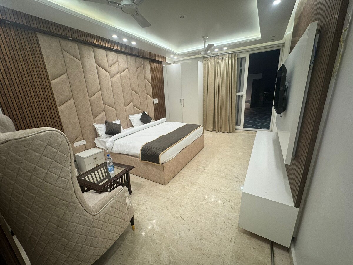 A spacious bedroom features a large bed with a neatly arranged duvet. A plush armchair sits next to a small wooden table. Light-colored walls are complemented by a textured headboard. A large window is dressed with light curtains, allowing gentle light to flow in.