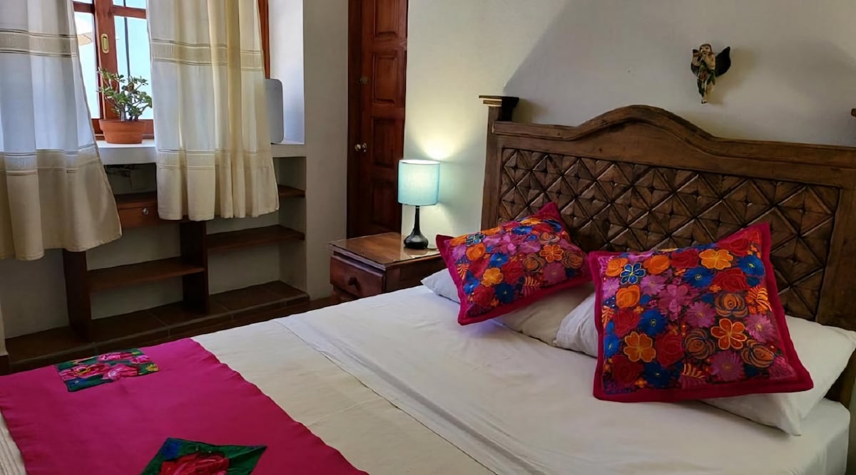 A cozy sleeping area features a wooden headboard with intricate carvings. Colorful pillows adorn a spacious bed, and natural light filters through curtains framing a window. A bedside lamp adds soft illumination, while a wooden cabinet and open shelves are visible in the background.