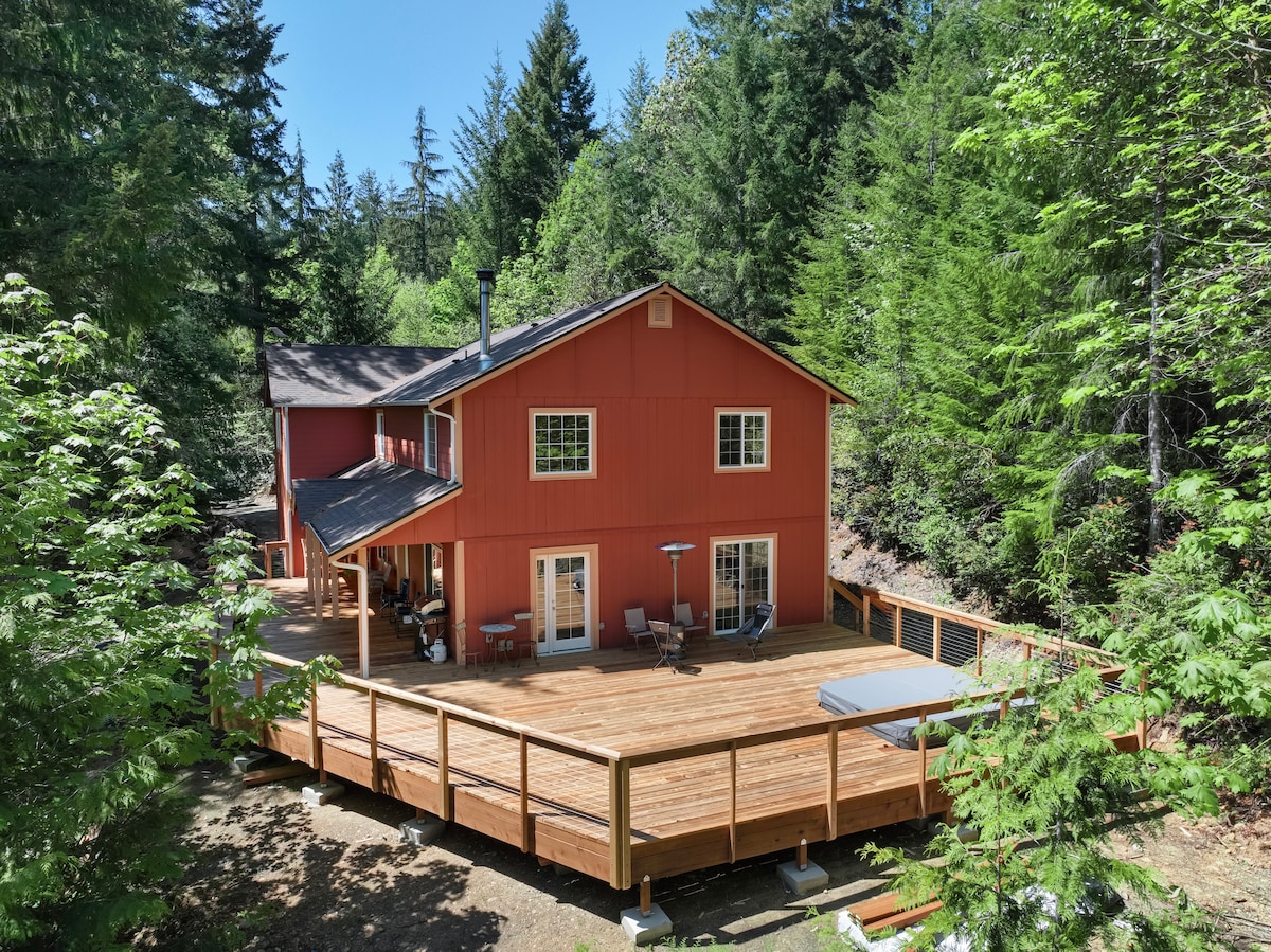The exterior of the retreat displays a large wooden deck surrounded by lush greenery. The modern red house features multiple large windows and a sloped roof. A hot tub is visible on the deck, accompanied by outdoor seating and a patio heater.