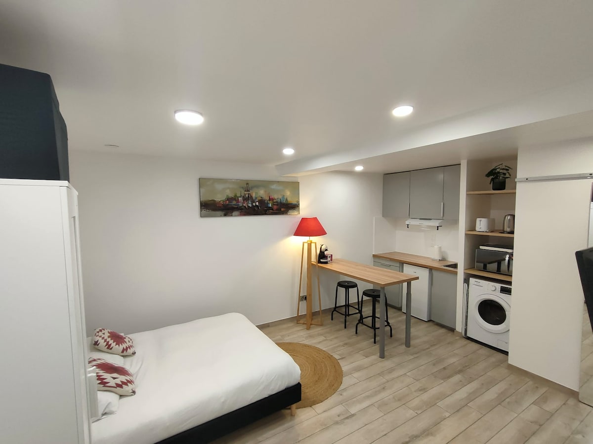 A compact living area features a spacious bed with white linens, complemented by patterned throw pillows. A small wooden table with two stools is positioned nearby. A washing machine and kitchen amenities are visible, while warm lighting from a lamp enhances the bright, neutral decor.