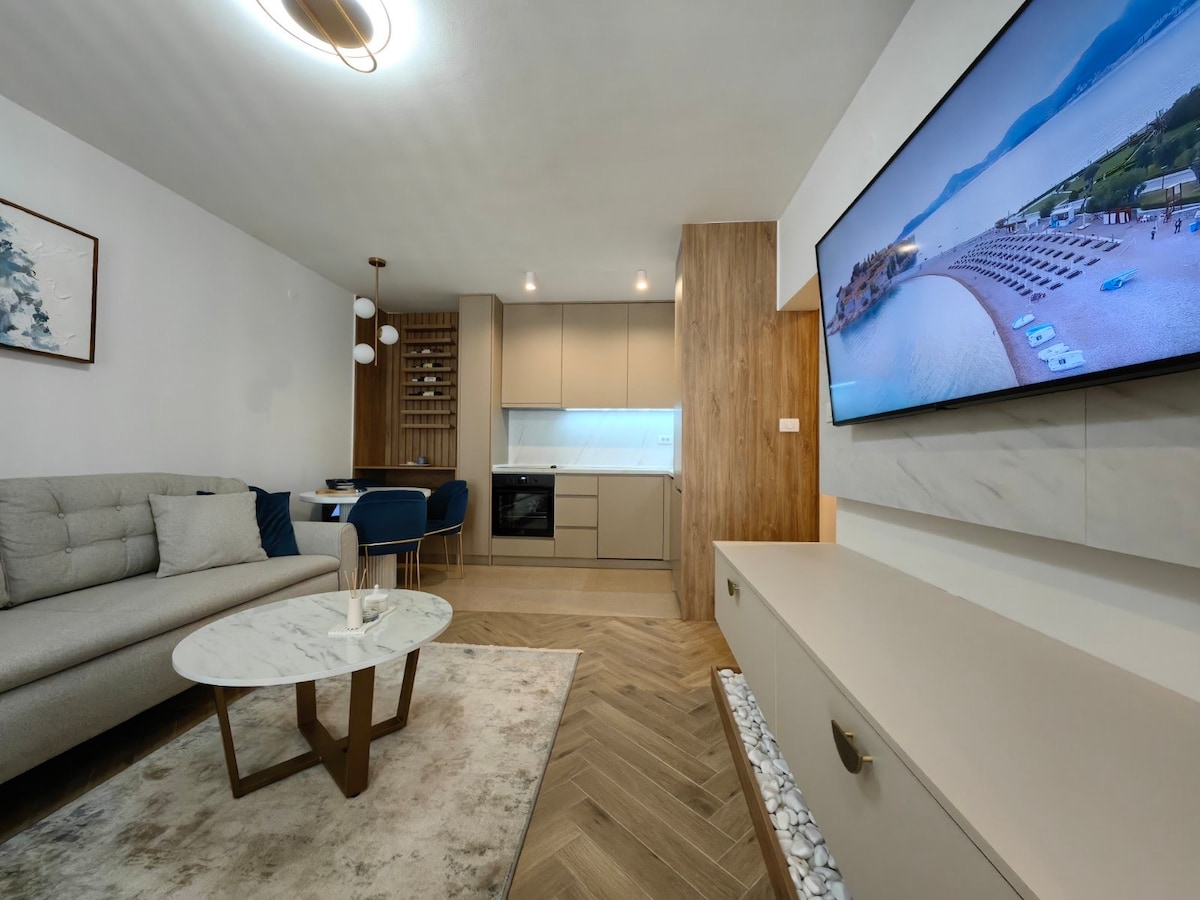 A modern living area is showcased, featuring a gray sofa paired with a marble coffee table. A flat-screen TV is mounted on a light-colored wall, oriented towards the seating. Natural light is filtered through a window, and a stylish dining table is positioned in the background.