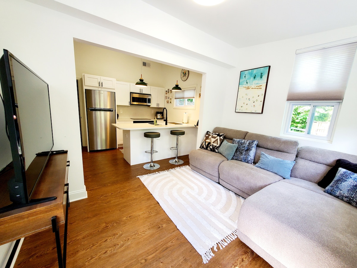 A cozy living area showcases a spacious sectional sofa adorned with decorative pillows. A flat-screen television is positioned on a wooden console. The adjoining kitchen features modern appliances and a small dining area with two bar stools. Natural light filters through nearby windows.