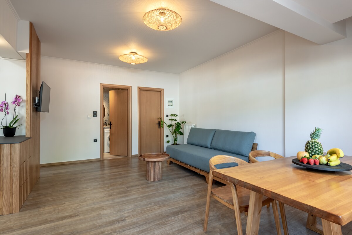 A modern living space features a comfortable sofa facing a wooden coffee table. A dining area is observed with a round table surrounded by chairs. Natural light enters through a nearby window, while decorative plants add a touch of greenery to the ambiance.