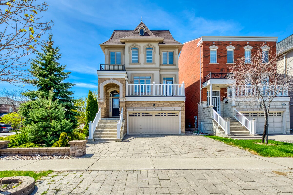 A grand three-story house with an elegant facade sits prominently among neighboring homes. Stone accents and large windows create an inviting look. A wide driveway leads to a two-car garage, and a spacious balcony is visible on the second floor, perfect for outdoor relaxation.