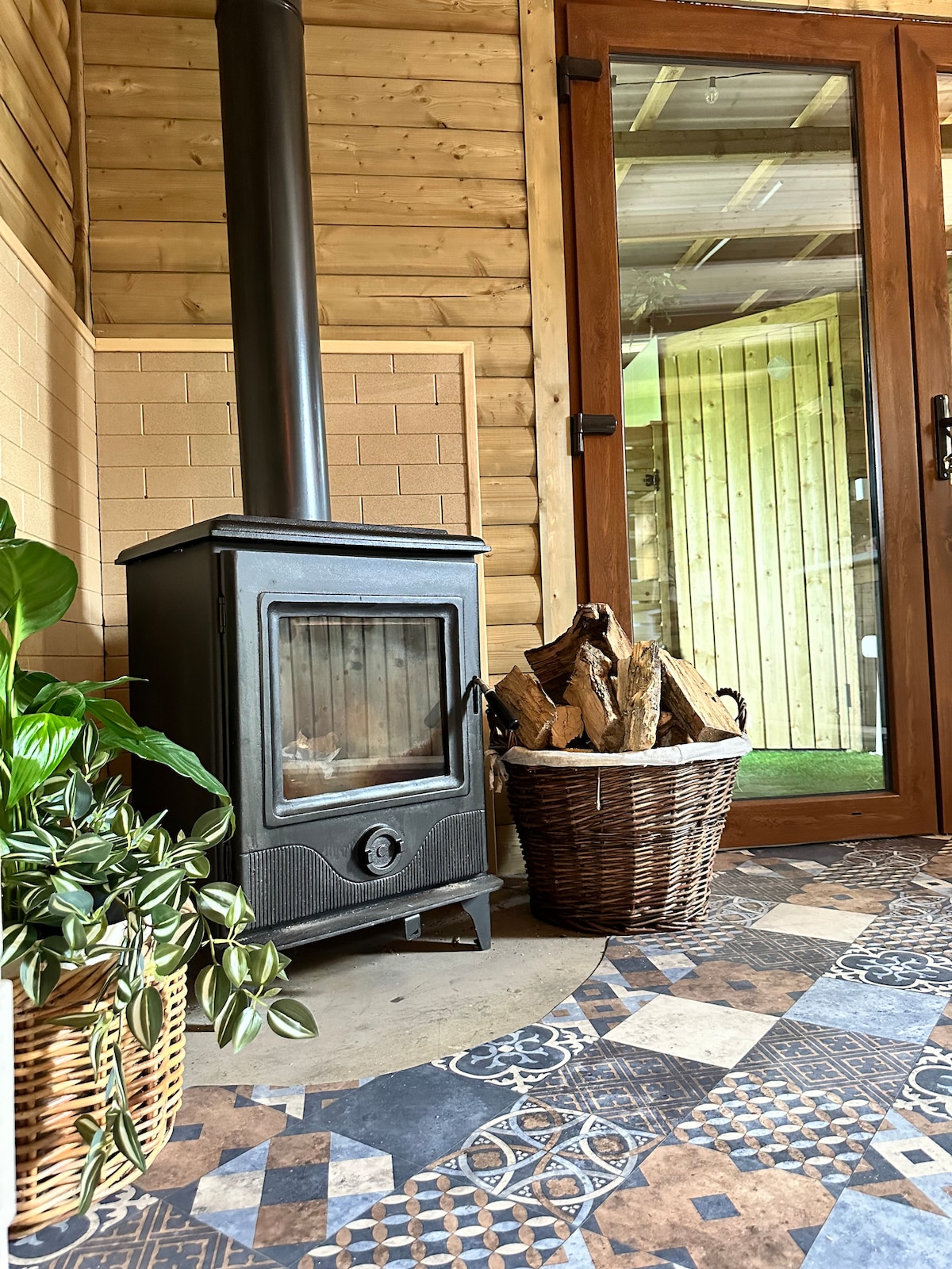 A cozy wood stove is positioned next to a rustic basket filled with firewood, enhancing the warmth of the space. Lush greenery is visible in a woven planter, with a patterned tile floor adding visual interest. Large glass doors provide access to the outdoors, with natural wood paneling surrounding the area.