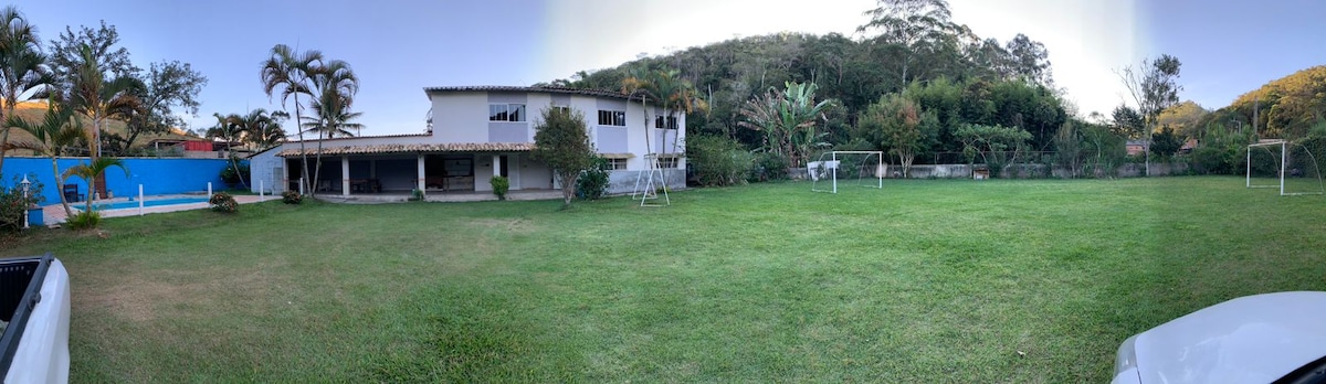 A panoramic view of the spacious outdoor area showcases a large green lawn with a soccer field in the foreground. The two-story house is seen on the left, with a covered patio extending outwards. Surrounding trees and plants create a natural backdrop.