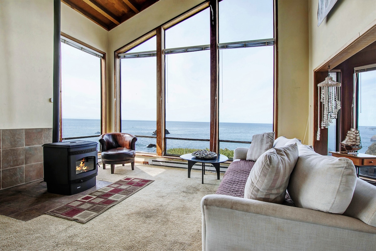 A spacious living area features a cozy couch and a comfortable chair, positioned near large windows that showcase expansive ocean views. Soft beige carpeting complements the warm tones of the wooden beams and a pellet stove serves as a focal point for relaxation.