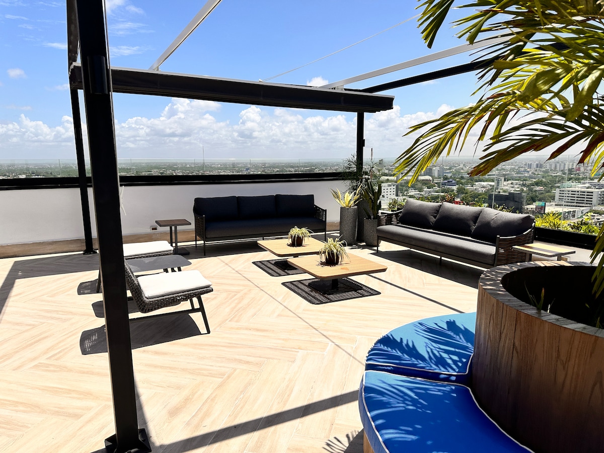 An outdoor terrace is showcased with a view of the surrounding city and sky. Comfortable seating options include a dark sofa and lounge chairs, complemented by potted plants. A wooden coffee table is positioned centrally, surrounded by decorative elements on the ground.