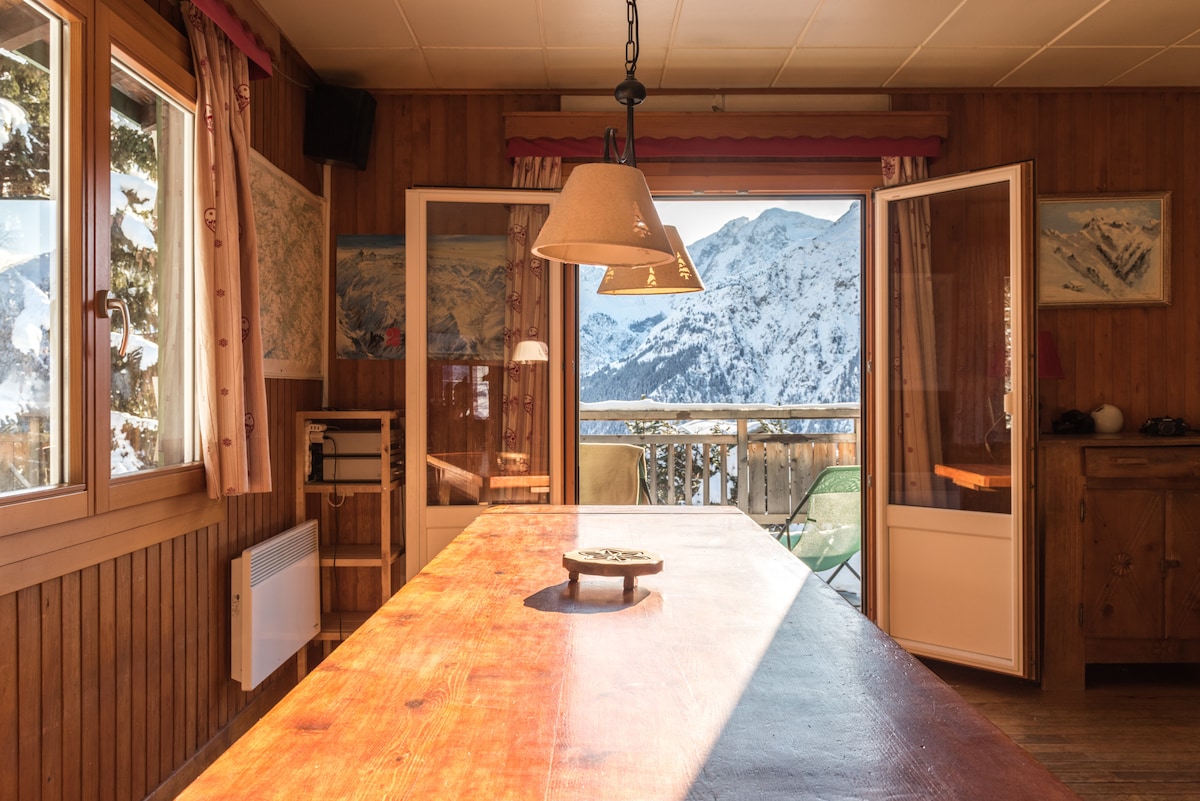 The dining area features a large wooden table illuminated by natural light from the open French doors. Mountain views are framed by the windows, and the rustic decor includes a pendant light and wall-mounted artwork.