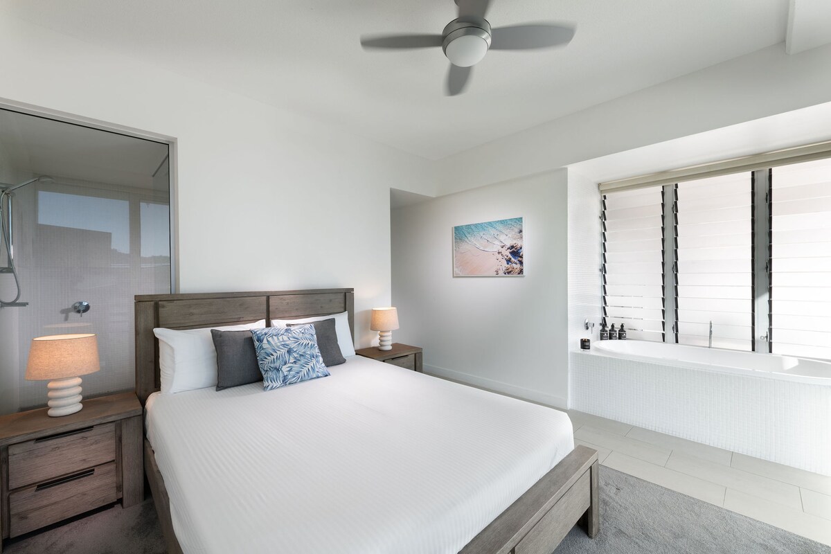 A bright and airy bedroom features a spacious bed with white linens and decorative pillows. Two bedside lamps provide soft lighting. A modern bathroom area is visible, with a glass shower. Natural light filters through the window, enhancing the serene atmosphere.