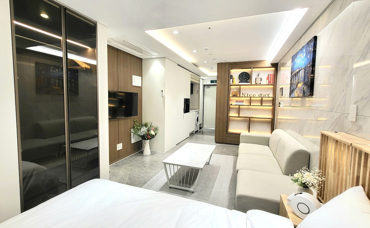 An open living space showcases a modern design, featuring a light-colored sofa, a coffee table, and a wall-mounted television. Soft lighting enhances the minimalist decor, while a flower arrangement adds a touch of greenery. Large windows allow natural light to brighten the room.